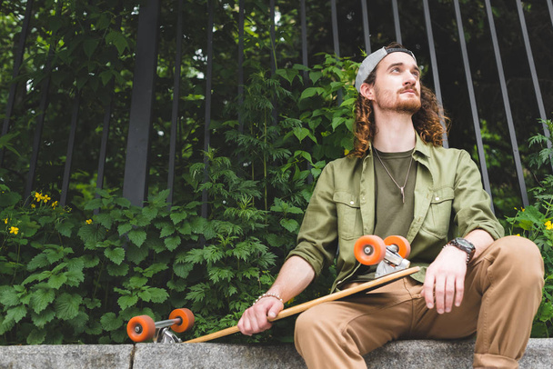 low angle view of handsome man sitting with skateboard on green plant background, looking away - Fotó, kép