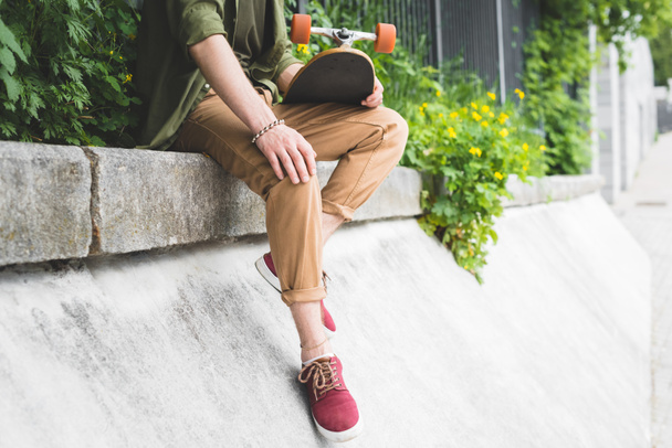 cropped view of man in casual wear holding skateboard in hand, sitting on border - Φωτογραφία, εικόνα