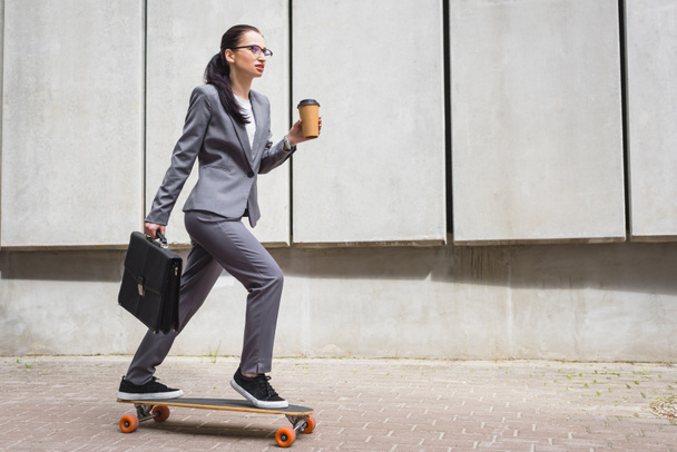 calm businesswoman in formal wear riding on skateboard, holding paper cup and briefcase in hands - Zdjęcie, obraz