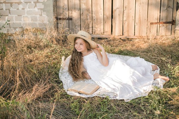 Cute little girl with blond long hair in a summer field at sunset with a white dress with a straw hat reading a book - Foto, Imagem