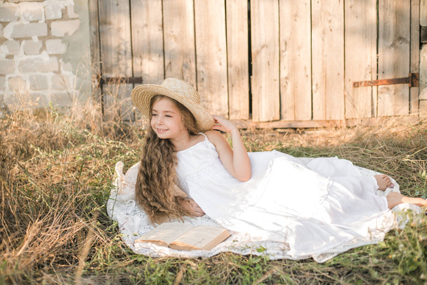 Cute little girl with blond long hair in a summer field at sunset with a white dress with a straw hat reading a book - Fotó, kép