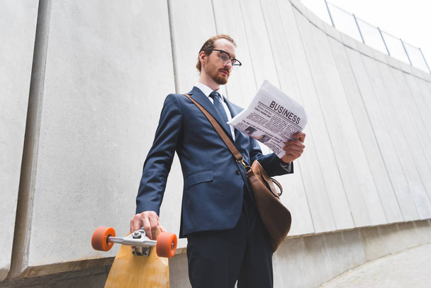low angle view of calm businessman in formal wear standing near skateboard, reading newspaper - 写真・画像