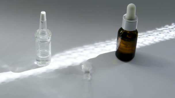 On the white surface are bottles with pipettes of dark and light glass with a cosmetic. The sun shines on them. Between them the glass rod is spinning and stopping indicates a white bottle - Footage, Video