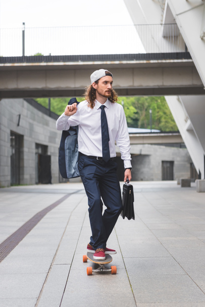 calm businessman with briefcase in hand riding on skateboard at street, looking away - Photo, Image