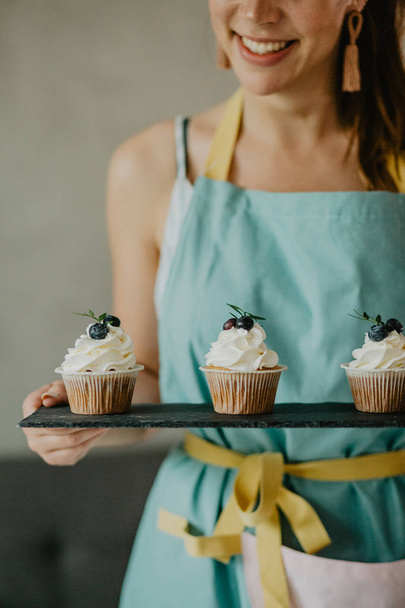 Beautiful woman in apron holding board with cupcakes - Foto, afbeelding