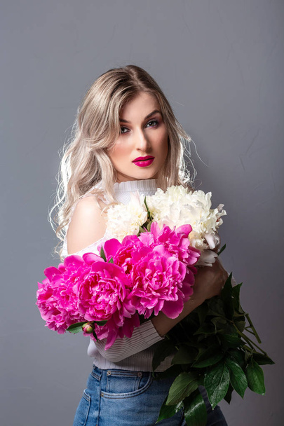 Dziewczyna z różowym bukietem piwonii, na szarym tle. Portret romantycznej blondynki z bukietem różowych kwiatów - Zdjęcie, obraz