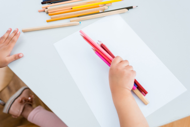 περικομμένη θέα του παιδιού κρατώντας πολύχρωμα μολύβια κοντά στο χαρτί  - Φωτογραφία, εικόνα