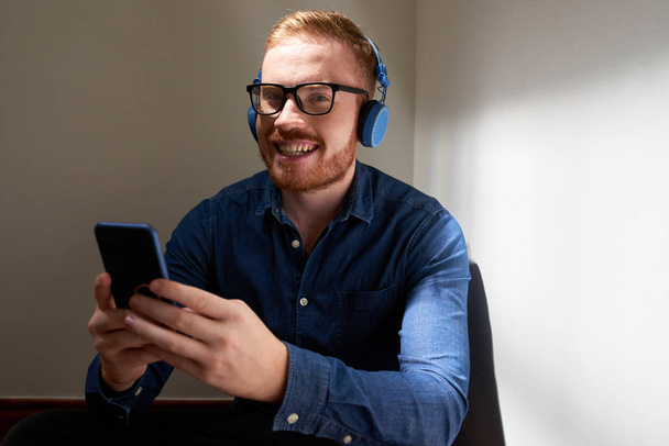 Portrait of handsome red haired young man in headphones using mobile app - Фото, зображення