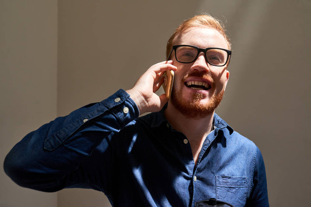 Portret van positieve vrolijke jonge man in glazen praten op telefoon - Foto, afbeelding