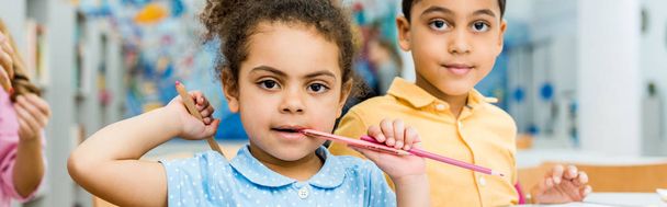 Panoramaaufnahme von süßen afrikanisch-amerikanischen Kind beißt Bleistift in der Nähe entzückendes Kind  - Foto, Bild
