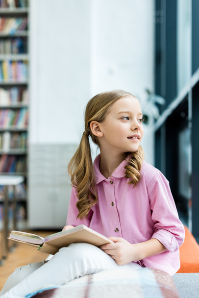 lindo y pensativo niño sentado y sosteniendo libro en la biblioteca
  - Foto, Imagen