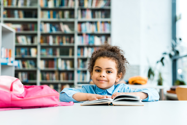 happy african american kid sitting and looking at camera near book and pink backpack  - Photo, Image