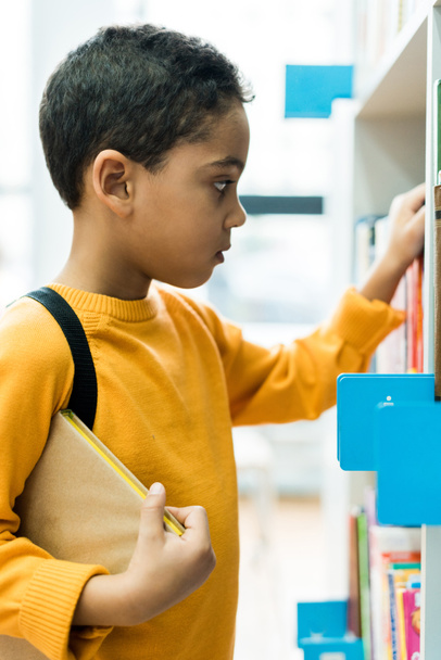 adorable african american boy standing and holding book in library  - 写真・画像