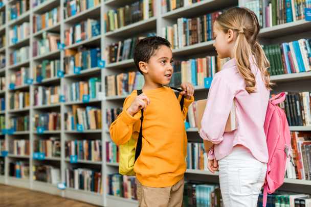 cute multicultural kids standing and talking in library  - Foto, Imagen