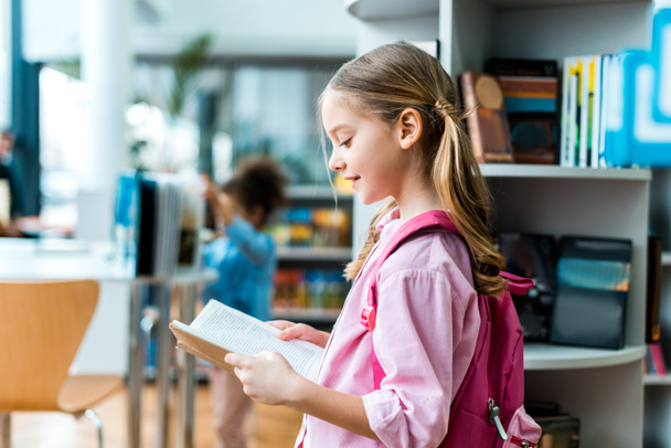 cheerful kid with pink backpack standing and reading book in library  - Photo, Image