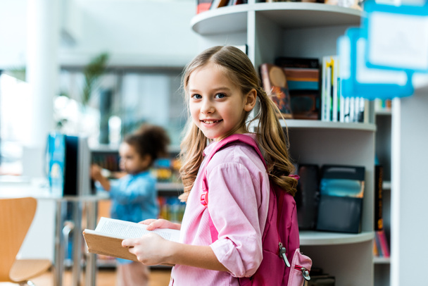 cheerful kid with pink backpack standing and holding book in library  - Фото, изображение