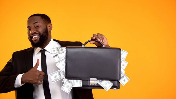 Joyful handsome black man in suit holding handbag with cash, showing thumbs-up - 写真・画像