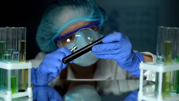 Scientist looking at black substance in tube with magnifying glass, examination - Photo, Image