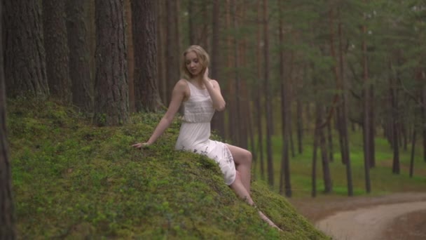 Piękna Młoda Blondynka siedzi w lesie nimfa w białej sukni w wiecznie zielonym drewnie - Materiał filmowy, wideo