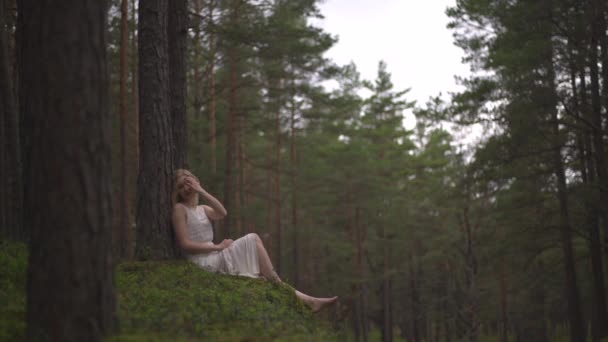Mooie jonge blonde vrouw zittend in bos nimf in witte jurk in groenblijvende hout - Video