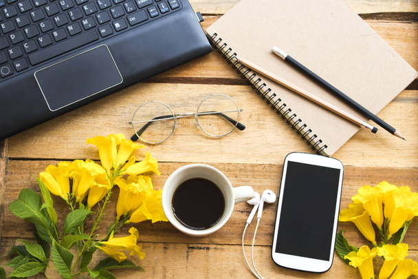Hober na notebook, mobilní telefon, počítač pro práci s horkou kávou, brýle a uspořádání žlutých květin plochý styl v kanceláři na pozadí dřevěné - Fotografie, Obrázek