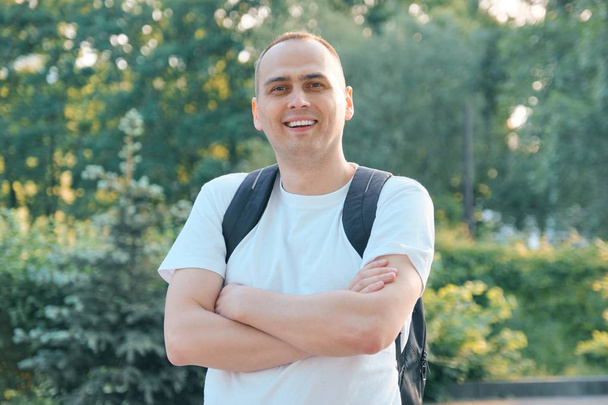 Outdoor portrait of mature smiling positive man. Confident male in white T-shirt with backpack with folded hands looking at the camera - Foto, imagen