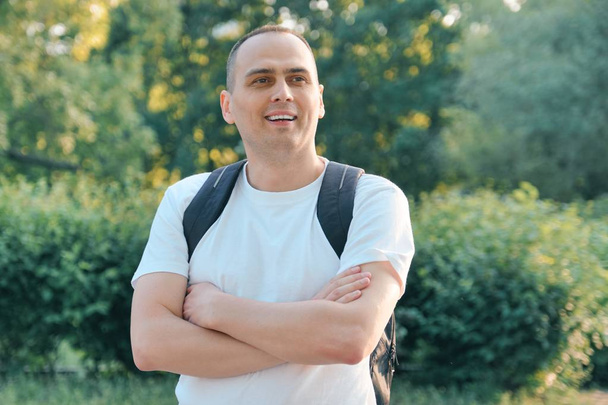 Outdoor portrait of mature smiling positive man. Confident male in white T-shirt with backpack with folded hands - Zdjęcie, obraz