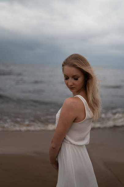 Fırtınalı rüzgar ve yağmur ile sıkıcı kasvetli havalarda dalgalar ile deniz kenarında beyaz elbise güzel genç sarışın kadın plaj perisi portresi kadar yakın - Fotoğraf, Görsel
