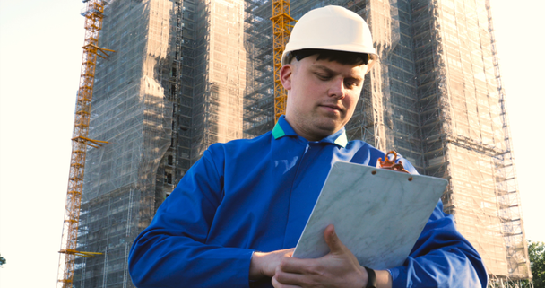 A young guy (male) architect, a businessman in a working suit, in a jacket with a folder with a tablet (mobile phone) in his hands checks the construction site and business building using various actions. - Séquence, vidéo