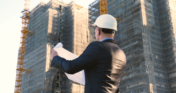A young guy (male) architect, a businessman in a working suit, in a jacket with a folder with a tablet (mobile phone) in his hands checks the construction site and business building using various actions. - Footage, Video