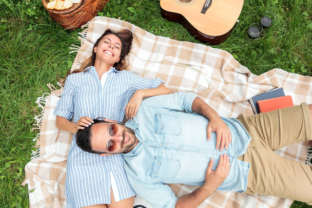 Ρομαντικό νεαρό ζευγάρι που χαλαρώνει σε ένα πάρκο. Ξαπλωμένοι στην πλάτη τους σε μια κουβέρτα πικ-νικ, αγκαλιές - Φωτογραφία, εικόνα