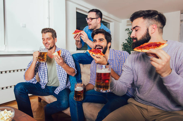 Группа из четырех друзей-мужчин пьет пиво и ест пиццу в доме
 - Фото, изображение