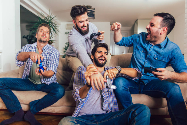 Улыбающиеся друзья-мужчины играют в видеоигры дома и веселятся. - Фото, изображение