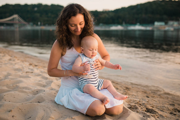 Young mother sitting on the beach with one year old baby son. Boy hugging, smiling, laughing, summer day. Happy childhood carefree game on the open sand, river lake, joy, fun. vacation concept - Photo, Image