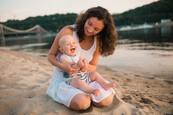 Young mother sitting on the beach with one year old baby son. Boy hugging, smiling, laughing, summer day. Happy childhood carefree game on the open sand, river lake, joy, fun. vacation concept - 写真・画像