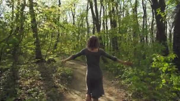 Young attractive woman in a gray dress wanders along pathway leading trough a thick forest in spring. Female wares sunglasses and enjoys nice weather. - Imágenes, Vídeo