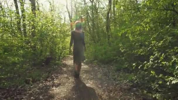 Young attractive woman in a gray dress wanders along pathway leading trough a thick forest in spring. Female wares sunglasses and enjoys nice weather. - Imágenes, Vídeo
