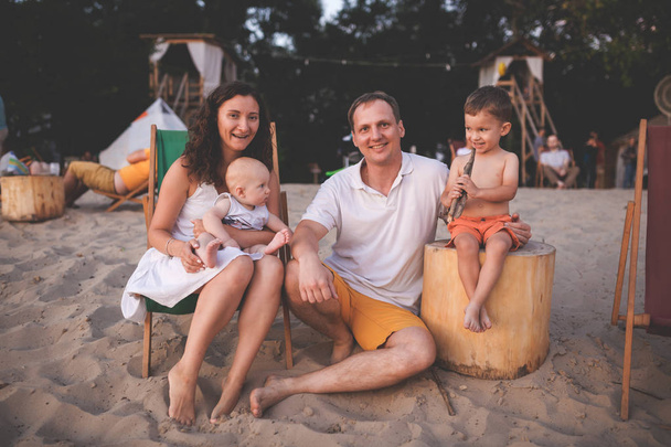 Mladá krásná kavkazská šťastná čtyřčlenná rodina sedící společně a objímající se při západu slunce v kavárně na pláži. Téma rodinná dovolená a rodinné hodnoty. Dva bratři kluci a máma, táta sedí na písečné pláži - Fotografie, Obrázek