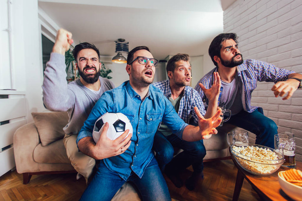 テレビで一緒にスポーツを観戦する男性たちが陽気に叫んで家にいる。Gr - 写真・画像