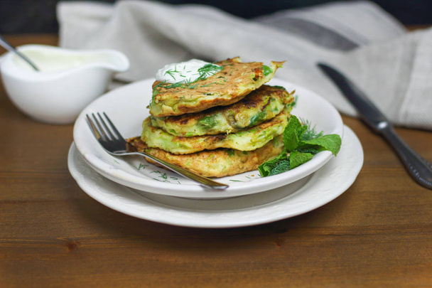 Proper nutrition, vegetarian breakfast gluten free,zucchini courgette pancakes with beans, mint on white plates with sour greek yougurt, black background, towel, fork, knife on wooden surface - Foto, afbeelding