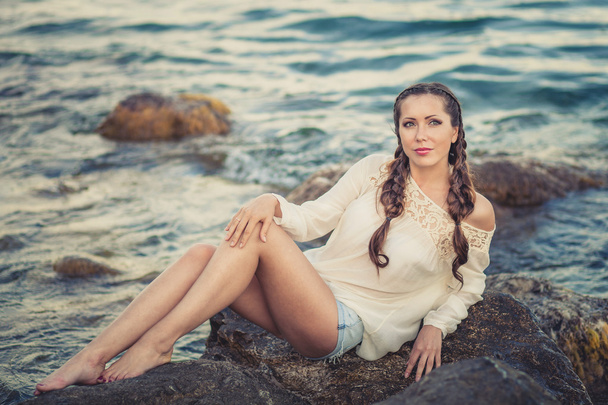 Beautiful young woman sitting on the stone on the seashore - Fotoğraf, Görsel