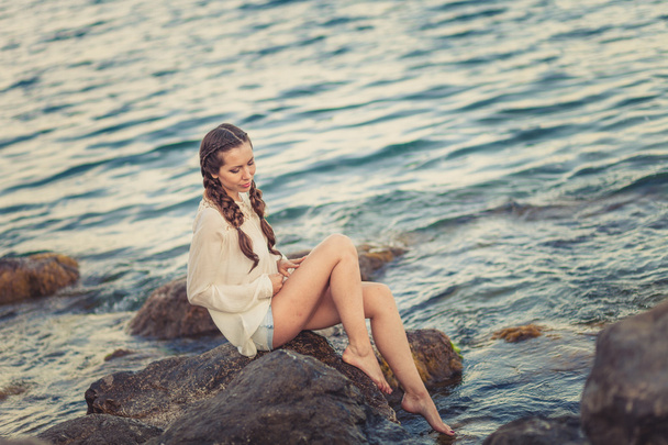 Beautiful young woman sitting on the stone on the seashore - Foto, Imagen