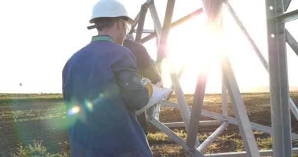 An electrician or builder and engineer, in a blue robe, in a yellow and white helmet, check the technical overview for strength, prepare electric poles, an electric tower, energy, and workers. - Video