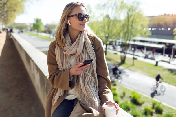 Mooie jonge vrouw luisteren naar muziek met draadloze koptelefoon en de smartphone in de straat. - Foto, afbeelding