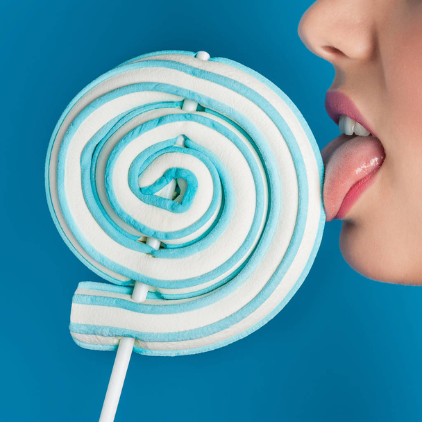 Woman licking lollipop close up image on blue background. - 写真・画像