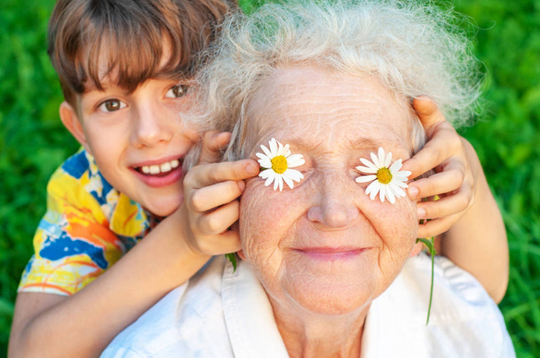 Der lachende Enkel mit seiner geliebten Großmutter verbringt seine Freizeit im Freien. legt der Junge seine Großmutter in die Augen der Kamille. Familienurlaub. - Foto, Bild