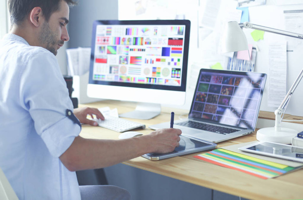 Portret van jonge ontwerper zittend in grafische studio achter laptop en computer terwijl hij online werkt. - Foto, afbeelding