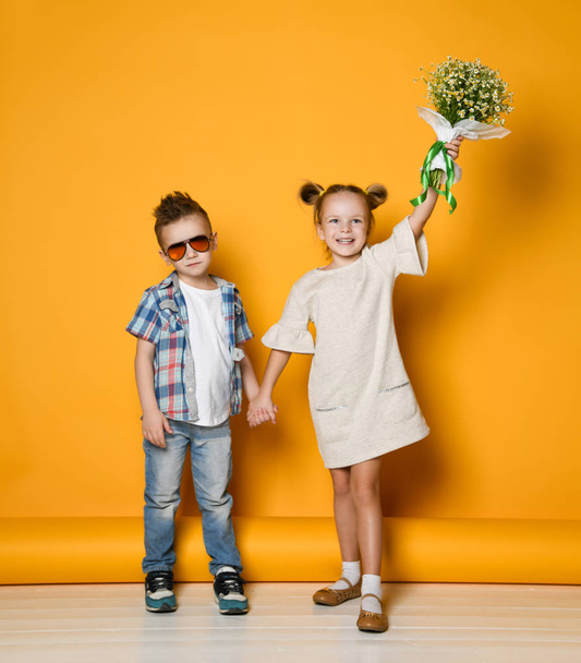 boy gave a bouquet of flowers to the girl. Children hold hands and look at the camera against the background of a yellow wall. - Фото, изображение