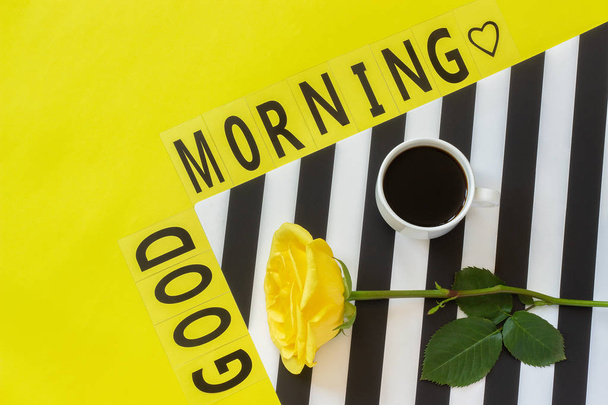 Text Good morning, Cup of coffee and yellow rose on stylish black and white napkin on yellow background. Minimal style. Concept good morning - Photo, Image
