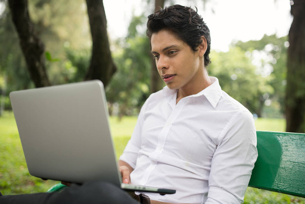 Young handsome man working with laptop on bench at park - Photo, image